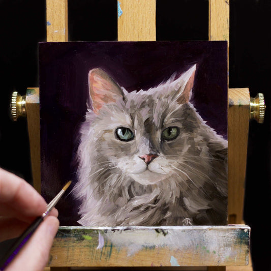 The painting of a green eyed, grey cat. The painting has a black- purple background sits on miniature easel