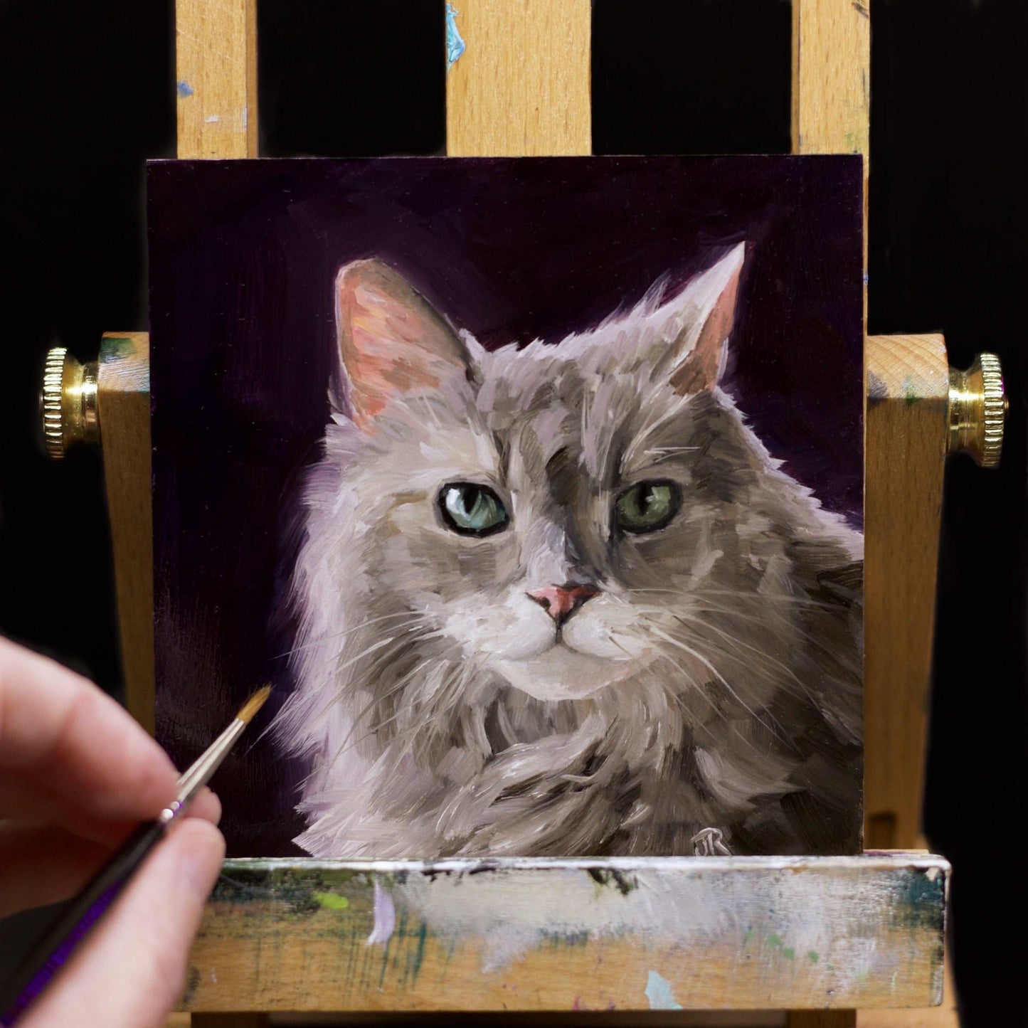 The painting of a green eyed, grey cat. The painting has a black- purple background sits on  miniature easel
